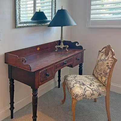 Sale Photo Thumbnail #206: Antique wooden writing desk paired with a toile-upholstered side chair, brass table lamp with a dark shade, and a mosaic-framed wall mirror.