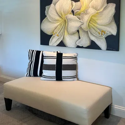 Sale Photo Thumbnail #209: Contemporary cream upholstered bench with dark wooden legs, topped with two black-and-white patterned accent pillows, positioned beneath a large canvas painting of white flowers on a dark background.