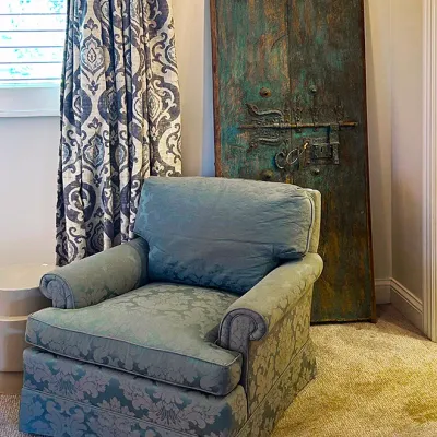 Sale Photo Thumbnail #236: Light blue damask armchair positioned in front of a pair of antique distressed wooden doors with ornate wrought iron hardware.