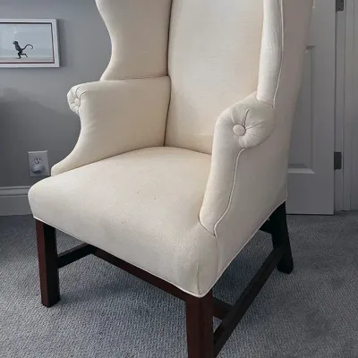 Sale Photo Thumbnail #20: Classic wingback armchair upholstered in cream fabric with button-tufted arm fronts and dark wooden legs.