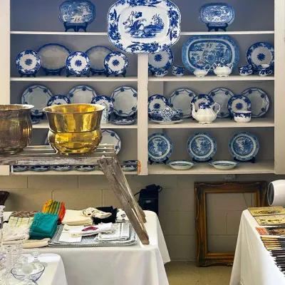 Sale Photo Thumbnail #134: Collection of vintage blue and white transferware porcelain, including platters, plates, and teapots, displayed on wooden shelving alongside polished brass planters and table linens.
