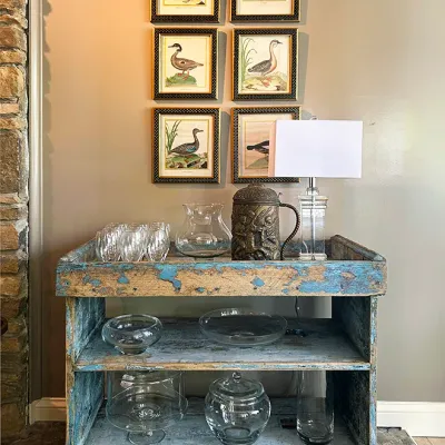 Sale Photo Thumbnail #114: Rustic distressed blue wooden three-shelf cart styled with glassware, metal steins, and a small table lamp, displayed beneath six framed bird art prints.