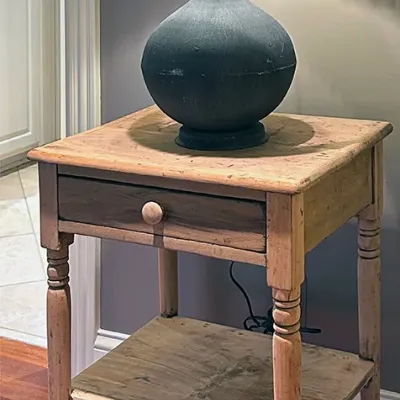 Sale Photo Thumbnail #235: Rustic pine side table with a single drawer and lower shelf, topped with an accent table lamp featuring a dark bulbous base and textured metallic shade.