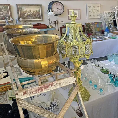 Sale Photo Thumbnail #280: Collection of home decor items featuring large brass planters, an ornate green and crystal table lamp, vintage glassware, linens, and framed artwork displayed on tables.