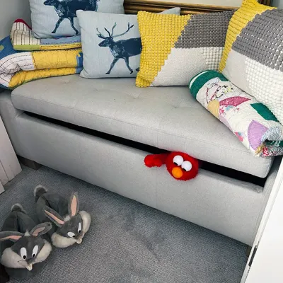 Sale Photo Thumbnail #213: Tufted light gray upholstered storage bench styled with throw pillows, folded quilts, an Elmo plush sticking out, and a pair of Bugs Bunny novelty slippers on the floor.