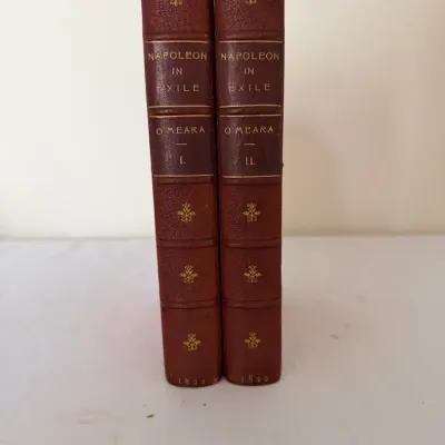 Sale Photo Thumbnail #82: Two-volume leather-bound set of 'Napoleon in Exile' by Barry E. O'Meara, dated 1822. The spines feature gilt lettering, decorative fleuron stamps, and raised bands.