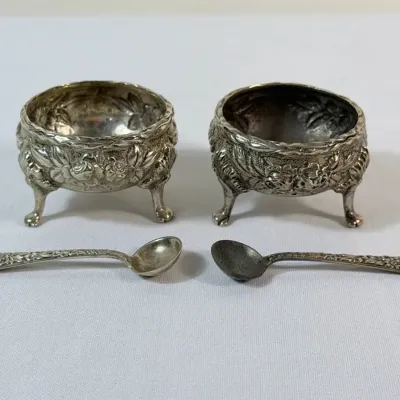Sale Photo Thumbnail #22: Pair of ornate silver-plated open salt cellars with three feet, floral repoussé detailing, and matching tiny spoons.