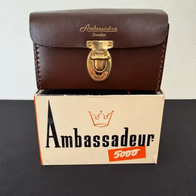Sale Photo Thumbnail #8: Vintage Ambassadeur 5000 fishing reel brown leather case stamped 'Ambassadeur Sweden' sitting atop its original box.