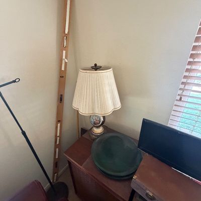 Sale Photo Thumbnail #133: Vintage ceramic table lamp with pleated fabric shade resting on a wooden cabinet, alongside a tall wooden level leaning against the wall.