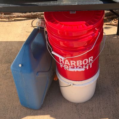 Sale Photo Thumbnail #264: Stack of 5-gallon plastic buckets, including red Harbor Freight buckets, alongside a blue plastic utility jug sitting beneath a table.