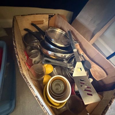 Sale Photo Thumbnail #81: Cardboard box filled with assorted cookware, including stainless steel pots and pans, a spatula, wooden spoons, and glassware.