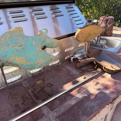 Sale Photo Thumbnail #70: Two decorative folk art metal fish sculptures on stands, displayed outdoors next to a stainless steel barbecue grill.