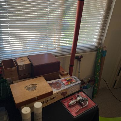 Sale Photo Thumbnail #191: Collection of tabletop items including cigar boxes, a boxed soprano ukulele, a packaged corkscrew, and spray cans set near a window.