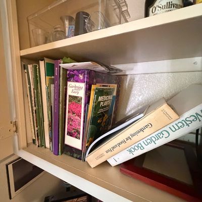 Sale Photo Thumbnail #76: Collection of vintage gardening books and guides arranged on a wooden cabinet shelf alongside barware and a clear acrylic container.