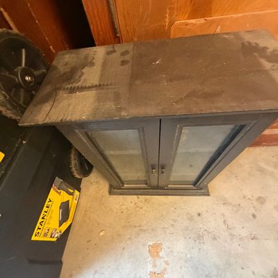 Sale Photo Thumbnail #214: Small black wooden storage cabinet with two glass-paneled doors, showing surface wear and dust.