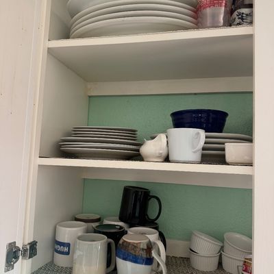 Sale Photo Thumbnail #18: Open kitchen cabinet filled with dishware, including stacks of white plates, ceramic mugs, plastic cups, and small ramekins.