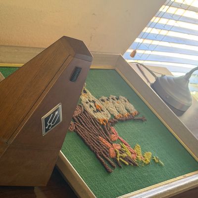 Sale Photo Thumbnail #36: Vintage Seth Thomas pyramid wood metronome resting beside a framed 1970s embroidered owl fiber art piece.
