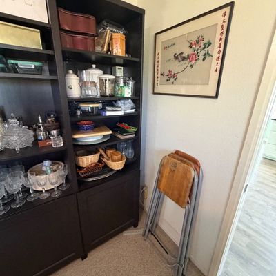 Sale Photo Thumbnail #152: Black wooden shelving unit filled with glassware and kitchenware alongside a set of folded metal snack TV tray tables and a framed Chinese silk embroidery hanging on the wall.
