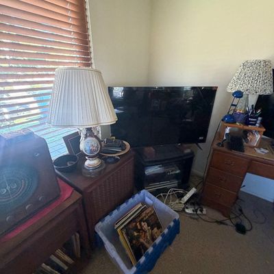 Sale Photo Thumbnail #130: Corner room layout with flat-screen television, vintage tabletop radio, table lamps, small wooden desk, and a plastic crate containing vinyl records.