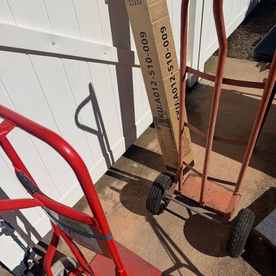 Sale Photo Thumbnail #265: Two red hand trucks with rubber wheels, positioned next to a tall cardboard shipping box outdoors.