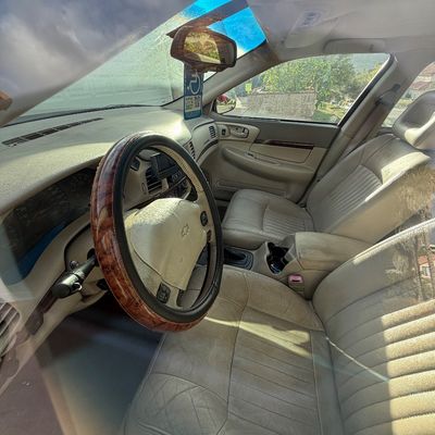 Sale Photo Thumbnail #10: Interior view of a late-1990s or early-2000s Chevrolet sedan featuring beige leather upholstery and a woodgrain-pattern steering wheel cover.
