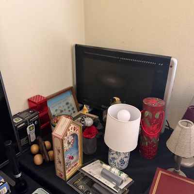 Sale Photo Thumbnail #193: A tabletop assortment of household items, including a small blue-and-white ceramic lamp, a decorative floral desk clock, framed artwork, and a flat-screen television.