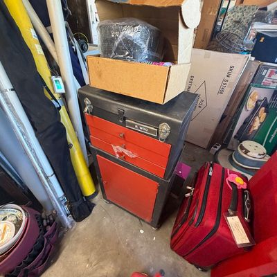 Sale Photo Thumbnail #212: Vintage Craftsman-style black and red metal tool chest on wheels, topped with a cardboard box, alongside a red rolling suitcase and gas cans in a garage setting.