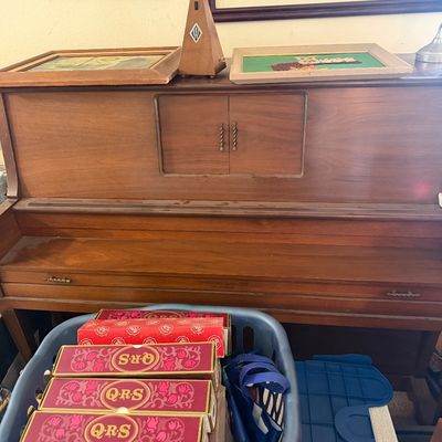 Sale Photo Thumbnail #163: Mid-century wooden upright player piano, accompanied by a basket containing boxed QRS player piano rolls and decorative artwork on top.