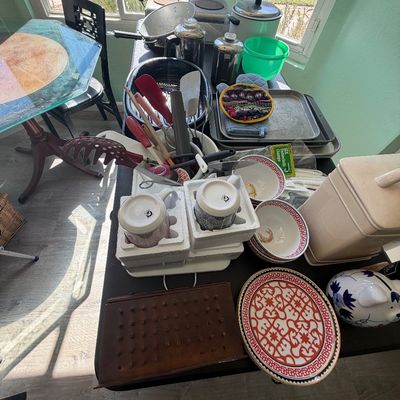 Sale Photo Thumbnail #87: Collection of assorted kitchenware and home items arranged on a folding table, including patterned serving dishes, mugs in foam packaging, cooking utensils, and small appliances.