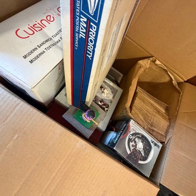 Sale Photo Thumbnail #213: Cardboard box packed with household items, including a boxed Cuisineart sandwich toaster, a Salton mini coffee grinder, picture frames, and paper bags.