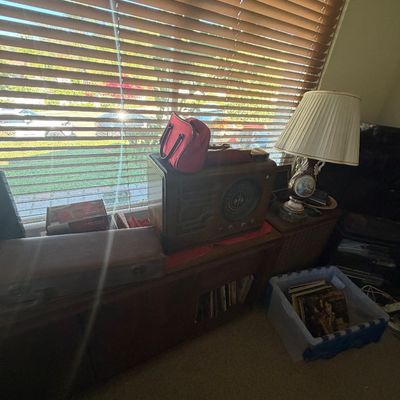 Sale Photo Thumbnail #131: Vintage tabletop tube radio sitting on a low wooden cabinet, surrounded by a leather hard instrument case, porcelain lamp, red handbag, and vinyl records in a crate.