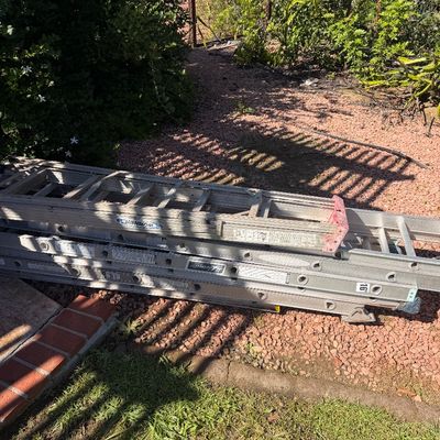 Sale Photo Thumbnail #66: Stack of aluminum extension ladders, including Werner and Louisville brands, lying outdoors on red gravel.