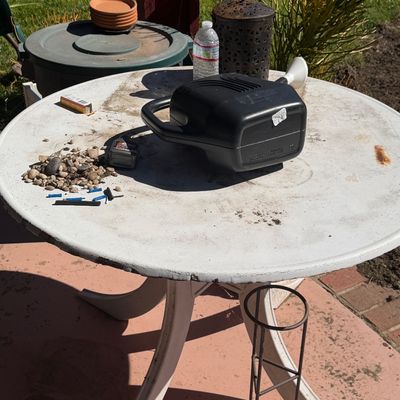 Sale Photo Thumbnail #63: White round outdoor patio table holding an assortment of garden and utility items, including a black plastic watering container, small pebbles, and decorative accessories.