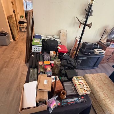 Sale Photo Thumbnail #122: Table displaying an assortment of household items, camera gear, small decor, and a boxed chef knife, alongside a standing halogen torchiere floor lamp.