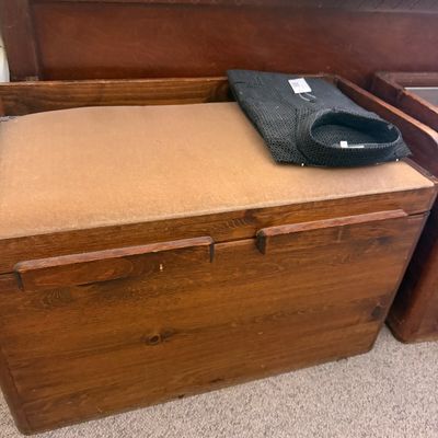 Sale Photo Thumbnail #294: Vintage wooden bench chest with a padded tan upholstered top, shown with a folded dark collared shirt resting on it.