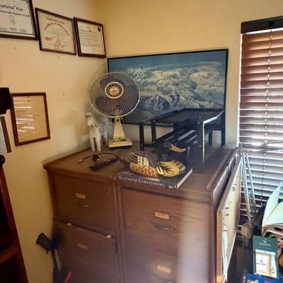 Sale Photo Thumbnail #116: Vintage wooden file cabinet topped with a decorative duck decoy, a vintage electric desk fan, a framed mountain print, and assorted home office decor.