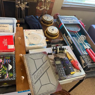 Sale Photo Thumbnail #137: Table array of miscellaneous household items including a vintage cash book, boxed 4-player badminton set, brass weather station, electronics, and fishing lure.