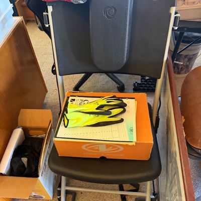 Sale Photo Thumbnail #166: Black ergonomically designed chair featuring an attached back support pad and a lower footrest frame, shown with a shoe box and work gloves on the seat.