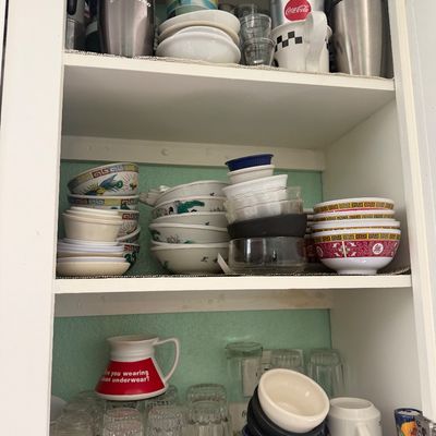 Sale Photo Thumbnail #19: White kitchen cabinet filled with assorted drinkware and dishware, including travel mugs, branded Coca-Cola and Starbucks items, novelty mugs, and traditional Chinese porcelain rice bowls.