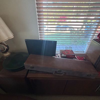 Sale Photo Thumbnail #132: Vintage long hard-shell musical instrument or equipment case resting on wooden furniture, alongside a computer monitor and table lamp.