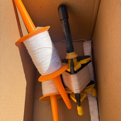 Sale Photo Thumbnail #272: Collection of kite string spools, including two orange plastic spools and one black and yellow winder, stored in a cardboard box.