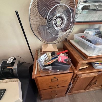 Sale Photo Thumbnail #97: Vintage oscillating electric table fan sitting atop a two-drawer wooden cabinet, alongside miscellaneous household items and electronics.