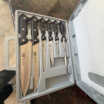 Sale Photo Thumbnail #88: A set of AJ kitchen knives with black handles, stored inside a molded gray carrying case alongside a honing steel.