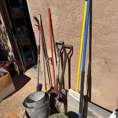 Sale Photo Thumbnail #255: Assortment of long-handled lawn and garden tools, galvanized metal buckets, and a vintage fuel can leaning against a stucco exterior wall.