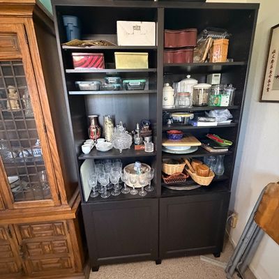 Sale Photo Thumbnail #151: Black wooden double bookcase unit with lower cabinets, filled with assorted glassware, kitchen storage tins, canisters, and decorative items.