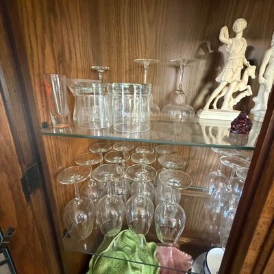 Sale Photo Thumbnail #146: A wooden hutch filled with assorted glassware including wine glasses and margarita glasses, classical-style figurines, and a green cabbage-leaf ceramic server.