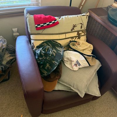 Sale Photo Thumbnail #136: Burgundy faux leather armchair holding an assortment of throw pillows and various hats, including bucket hats and a beanie.