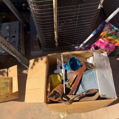 Sale Photo Thumbnail #253: Cardboard box filled with assorted household goods and tools, including leather belts, a blue funnel, and leather sheaths, next to an outdoor Lennox AC unit.