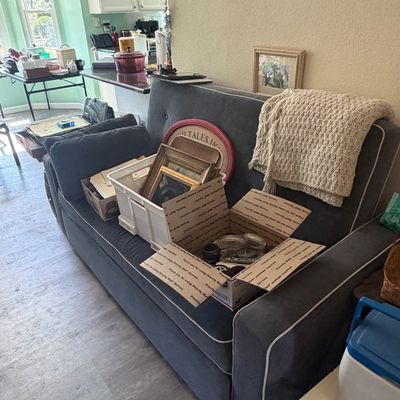 Sale Photo Thumbnail #109: Contemporary dark gray upholstered sofa with white piping, holding various household goods, framed artwork, and storage boxes.