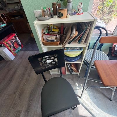 Sale Photo Thumbnail #82: Contemporary black wooden dining chair featuring a geometric cutout backrest and vinyl seat, positioned in front of a white shelving unit.